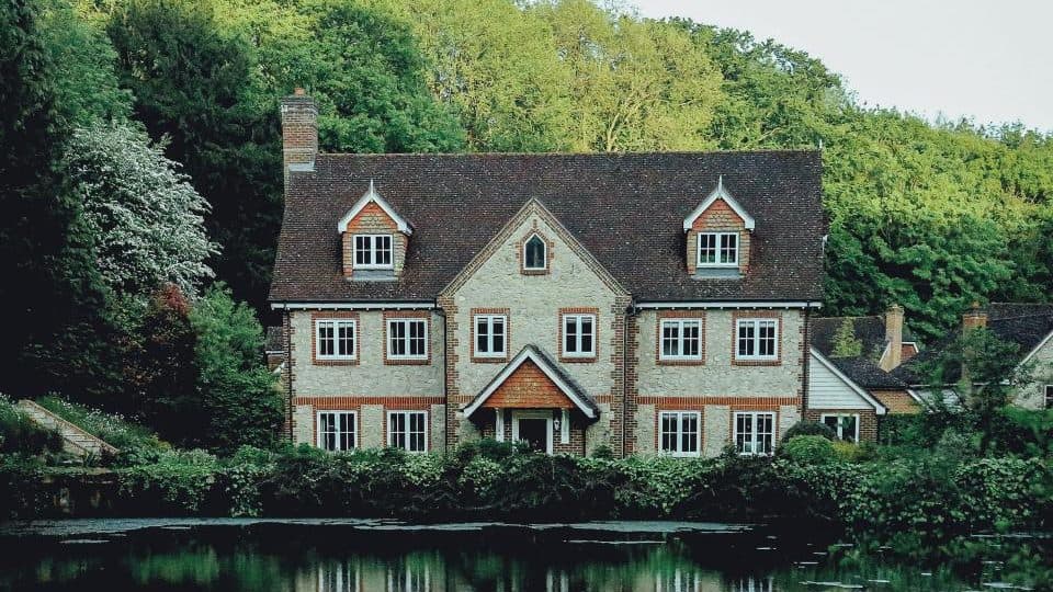Large stone house with dormer windows among trees beside a pond